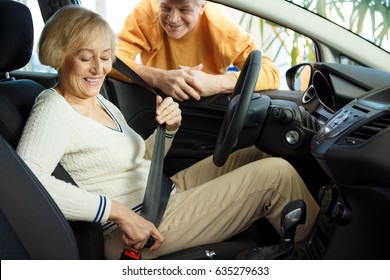 Senior Woman Sitting In A New Car Fastening Seatbelt Smiling Joyfully Her Loving Husband Standing Near Travelling Automotive Transportation Vehicle Renting Rental Service Driving Safety Insurance