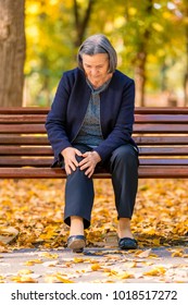 Senior Woman Sitting On Bench In Autumn Park And Having Knee Pain. Arthritis Pain Concept. Focus On Her Face.