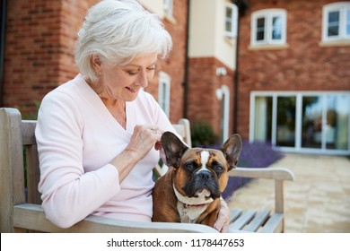 Senior Woman Sitting On Bench With Pet French Bulldog In Assisted Living Facility