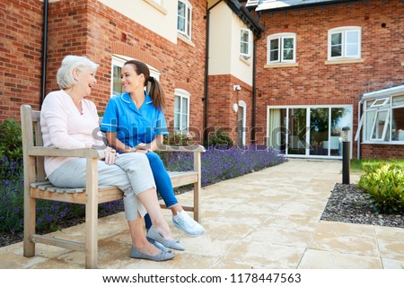 Senior Woman Sitting On Bench And Talking With Nurse In Retirement Home