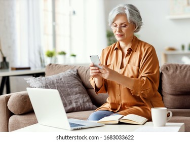 Senior Woman Sitting On Couch Near Table With Notepad And Laptop And Browsing Smartphone While Working On Remote Project At Home
