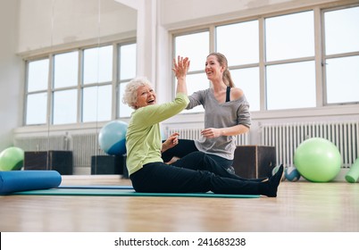 Senior Woman Sitting On Fitness Mat Giving High Five To Her Personal Trainer At Gym. Excited Old Woman Rejoicing Health Success With Her Instructor At Rehab.