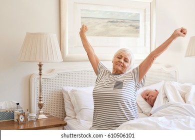 Senior woman sitting on her bed in the morning yawning with arms raised in a stretch with her husband sleeping next to her  - Powered by Shutterstock