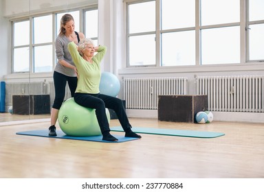 Senior Woman Sitting On A Pilates Ball  Exercising At Health Club Being Assisted By Her Personal Trainer. Physical Therapist Helping Senior Woman In Her Workout At Gym.