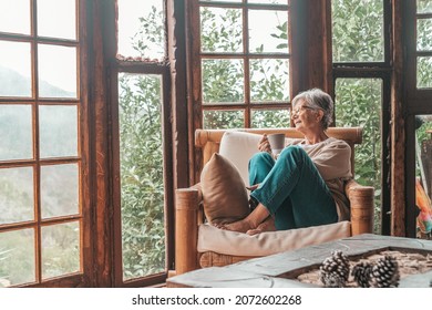 Senior woman sitting on sofa and holding coffee mug while looking away at home. Elderly woman relaxing on couch in living room. Thoughtful woman having coffee on sofa at home