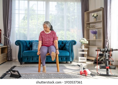 Senior woman sitting on a wooden chair, raising leg to stretch muscles and knees, Training exercise online with tablet In Living Room During Quarantine