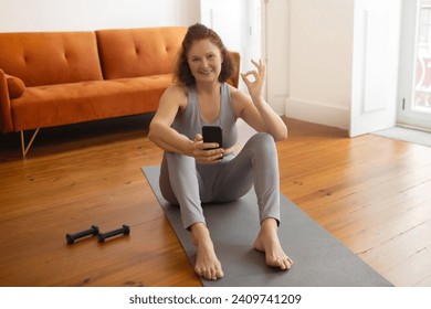 Senior woman sitting on yoga mat and showing OK sign while holding smartphone, happy elderly lady indicating satisfying workout session in her home, recommending fitness tracker application - Powered by Shutterstock