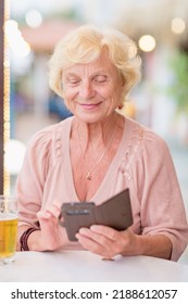 Senior woman sitting at a table in a summer cafe, looking at smartphone and drinking beer from a tall glass