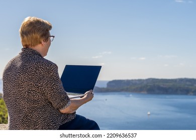 Senior Woman Sitting Using A Laptop In Front Of A Seascape, On A Sunny Day