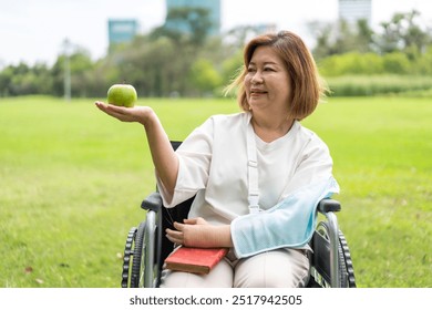 Senior woman sitting in a wheelchair holding a green apple in the park, enjoying healthy lifestyle, relaxation, recovery, outdoor activity, active elderly, independence, positivity, fresh air, smiling - Powered by Shutterstock