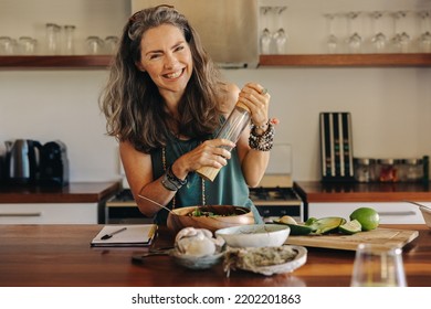 Senior woman smiling at the camera while preparing some plant-based food. Cheerful woman following a healthy vegan recipe in her kitchen. Happy mature woman eating clean at home. - Powered by Shutterstock