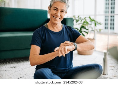 Senior woman smiling at the camera while checking her heart rate on a smartwatch. Mature woman monitoring her progress to stay on track with her fitness goals as she works out with yoga at home. - Powered by Shutterstock