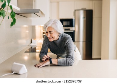 Senior woman smiling happily while using smart devices in her kitchen. Cheerful elderly woman using a home assistant to perform tasks at home. - Powered by Shutterstock