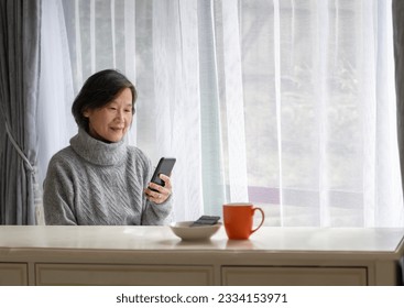 Senior woman smiling and reading smartphone. A coffee cup and dark chocolate on white table.   - Powered by Shutterstock