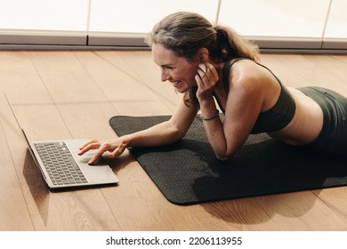 Senior Woman Smiling While Joining An Online Fitness Class On A Laptop. Happy Woman Following An Online Yoga Tutorial At Home. Cheerful Senior Woman Lying On An Exercise Mat.