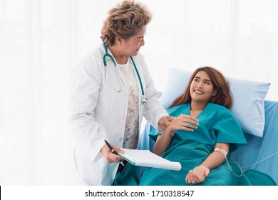 The senior woman specialist doctor with stethoscope checking up happy young Asian female patient lying in bed with receiving an intravenous saline drip in recovery room. Healthcare, medical insurance. - Powered by Shutterstock