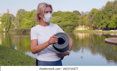 Senior Woman In Sportswear And Medical Mask Holding Fitness Mat Standing Near River In Summer Park. Active Mature Female In Safety Mask Resting After Yoga Class Outdoors