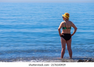 Senior Woman Standing Alone In Front Of The Mediterranean Sea At A Beach In A Sunny Summer Day. Older Female Enjoying Retirement. Full Body Length Shot.. Back View.