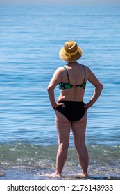 Senior Woman Standing Alone In Front Of The Mediterranean Sea At A Beach In A Sunny Summer Day. Older Female Enjoying Retirement. Full Body Length Shot.. Back View.