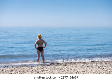 Senior Woman Standing Alone In Front Of The Mediterranean Sea At A Beach In A Sunny Summer Day. Older Female Enjoying Retirement. Full Body Length Shot.. Back View.