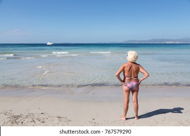 Senior Woman Standing Alone In Front Of The Sea / Ocean At A Beach In A Sunny Summer Day. Older Female Enjoying Retirement. Winter Getaway Concept. Back View. Full Body Length Shot.