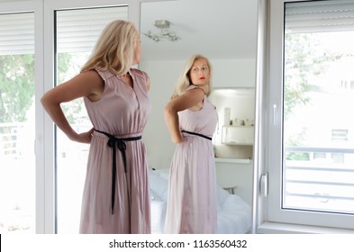 Senior Woman Standing In Front Of Mirror Wearing A Long Pink Dress.