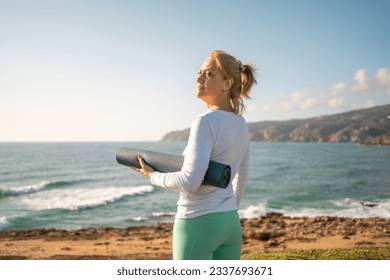 Senior woman standing ocean shore, holding yoga mat. She appears to be happy and enjoying the scenic views before her yoga session. This depicts a harmonious and healthy senior lifestyle concept. - Powered by Shutterstock