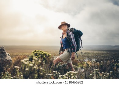 Senior woman standing on a hill wearing a backpack and looking away. Adventure seeking woman trekking through the bushes on a hill.