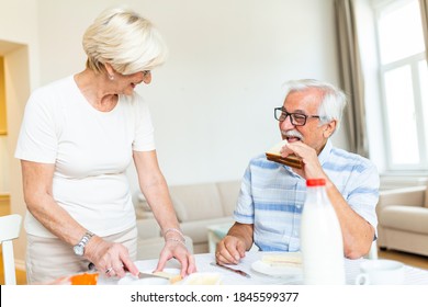 Senior Woman Standing And Preparing Breakfast. Elderly Couple Feeling Happy While Having Breakfast At Home. Old Man Eating Sandwich Made By His Lovely Wife. Couple Smiling