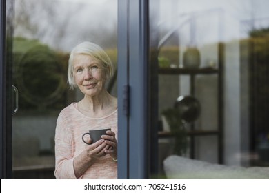 Senior woman is standing at the window in her home, enjoying a cup of tea. - Powered by Shutterstock