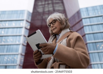 Senior woman in stylish coat reading tablet outside modern glass building during late afternoon - Powered by Shutterstock