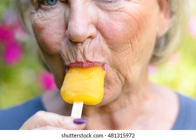 Senior Woman Sucking On An Iced Orange Lolly Outdoors On A Summer Day Cooling Off From The Heat, Close Up Cropped View On Her Mouth And Lips