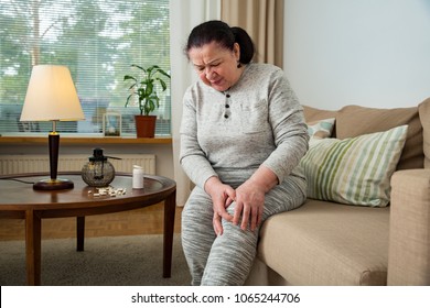 Senior Woman Suffering From Pain In Knees At Home. Holding Her Knee And Massaging With Hands, Feeling Exhausted, Sitting On Sofa In Living Room. Expressing Pain On Face. Medications And Pills On Table