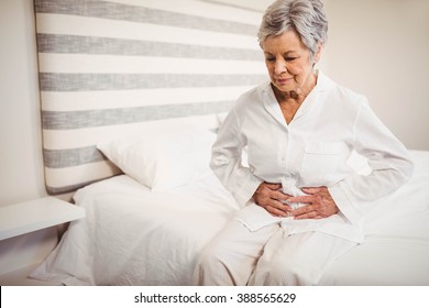 Senior woman suffering from stomach ache sitting on bed in bedroom - Powered by Shutterstock