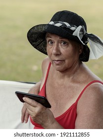 Senior Woman In A Summer Park, Holding Mobile Phone And Looking At Camera. She Is Communicating Online With Friends Or Family, Reading News, Looking 