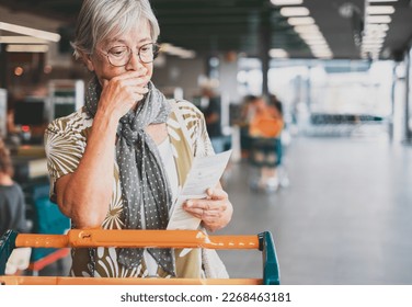 Senior woman in the supermarket checks her grocery receipt looking worried about rising costs - elderly lady pushing shopping cart, consumerism concept, rising prices, inflation - Powered by Shutterstock