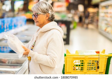 A Senior Woman At The Supermarket In Froozen Food Department With Shopping Cart Reading Carefully The Label On A Product Before To Buy It. Active Elderly People Everyday Life Concept