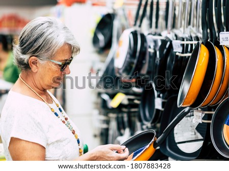 Senior woman at supermarket in houseware department chooses a frying pan. Active elderly person. Consumerism concept