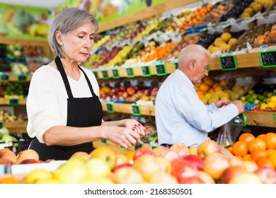 Senior Woman Supermarket Worker Putting Fruits On Shelves. Old Man Shopping In Background.