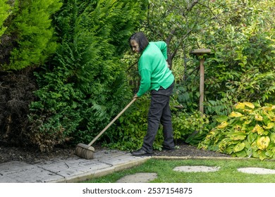 Senior woman sweeping garden pathway with broom, surrounded by lush green foliage, performing outdoor cleaning in backyard garden - Powered by Shutterstock