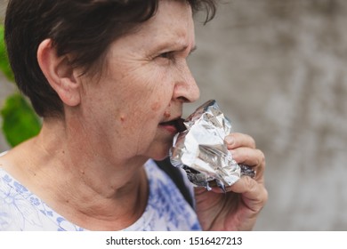 Senior Woman Taking A Bite From A Sweet Chocolate Bar Outdoors - Cute Old Lady Having A Quick Tasty Snack - Joyful And Happy Elderly Person With Healthy Eating Habits