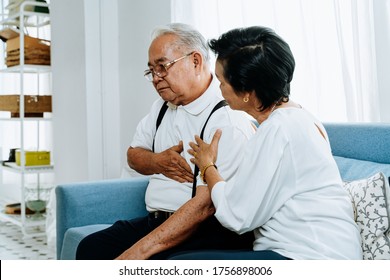 Senior Woman Taking Care Of Elderly Asian Man Suffering From Heartburn While Sitting On Sofa In Cozy Living Room At Home. Old Pensioner Sick Patient