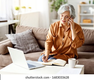 Senior Woman Talking With Client On Smartphone And Writing In Notepad While Sitting On Sofa And Working On Remote Project At Home
