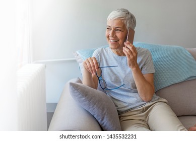 Senior Woman Talking On Her Mobile Phone. Senior Woman Has A Happy Conversation At Cellphone. Smiling Senior Woman Using Phone Sitting On Couch At Home.