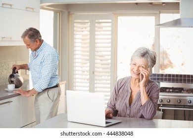 Senior Woman Talking On Mobile Phone With Husband Making Tea In Background At Home