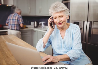 Senior woman talking on phone while using laptop and man working in kitchen at home