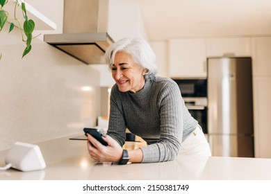 Senior Woman Talking To A Smart Speaker In Her Kitchen. Happy Elderly Woman Using A Virtual Assistant To Perform Household Tasks In Her Smart Home.