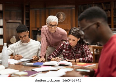 Senior Woman Teacher Working With College Students In Library. Graduates Consulting With Their Lecturer Informally During A Break In Library. Group Of Students Studying With Their Professor.