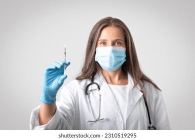 Senior woman therapist in white coat and protective mask showing syringe with vaccine, isolated on gray background, selective focus. Doctor doing vaccination, immunization for health care - Shutterstock ID 2310519295
