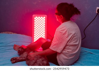 Senior woman treating sore leg with the red light therapy panel and wearing eye protection while petting a cat at home on the bed - Powered by Shutterstock